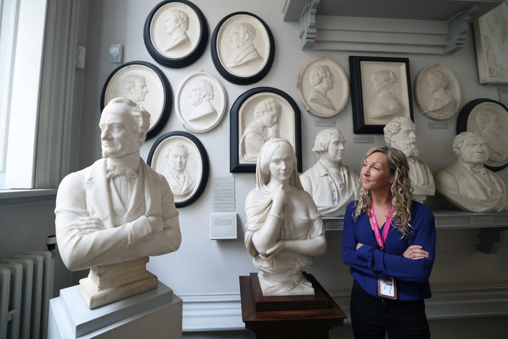 photo of Alex standing crossed arms in a gallery interior, the space lined with plaster reliefs, white sculpted busts on plinths to the left and foreground