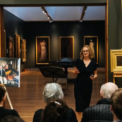 view of a darkened art gallery interior, seated audience members to the foreground, we can see the backs of their heads; the speaker before them gesturing, facing the camera, pictures in gilt frames in the room behind