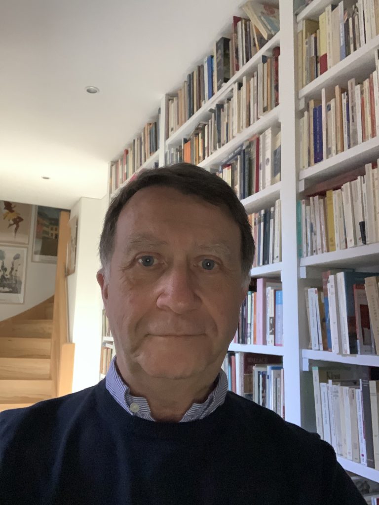 selfie-like photo image of Frederic, head and shoulders in an interior, filled bookshelves behind to the right, an open doorway with stairs ascending at the rear of the room