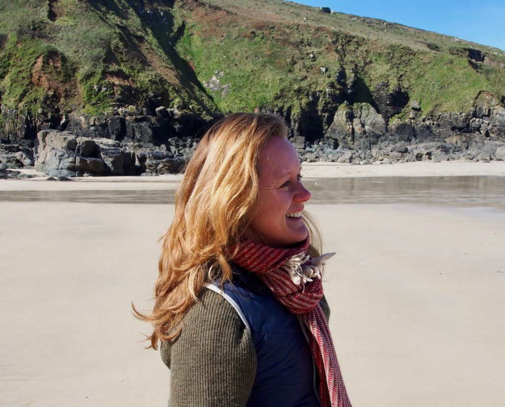 portrait photo of Lara, in profile facing to right, on a beach, green cliff behind, sunlit