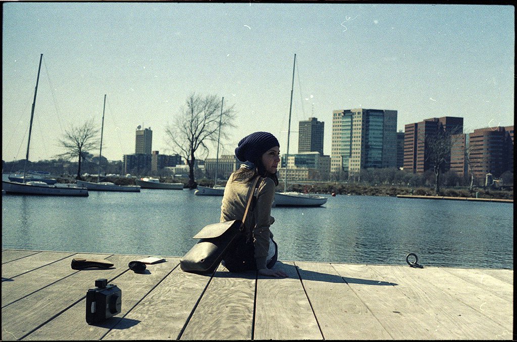 photographic, exterior view of a harbour, tall modern buildings and tree in distance, a wooden decking surface nearest to us, we can see Laura from behind, sitting with legs over the edge, looking over their shoulder