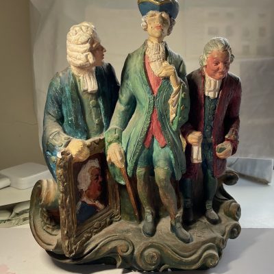photo of a realistically painted plaster sculpture, of three eighteenth century figures, their features somewhat caricatured,