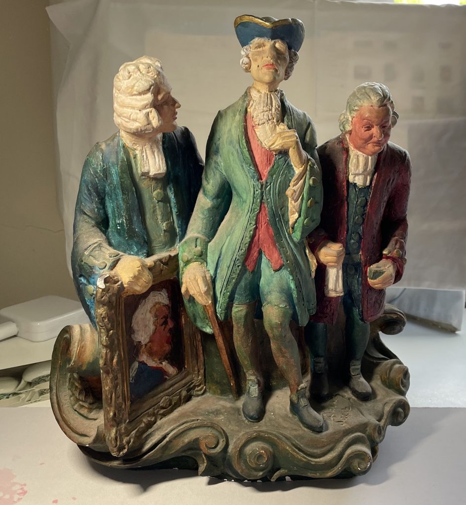 photo of a realistically painted plaster sculpture, of three eighteenth century figures, their features somewhat caricatured,