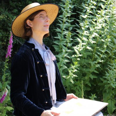 portrait photo of Grace, seated, in daylight, with widebrimmed hat and opened book on knee, looking upwards, large plants behind