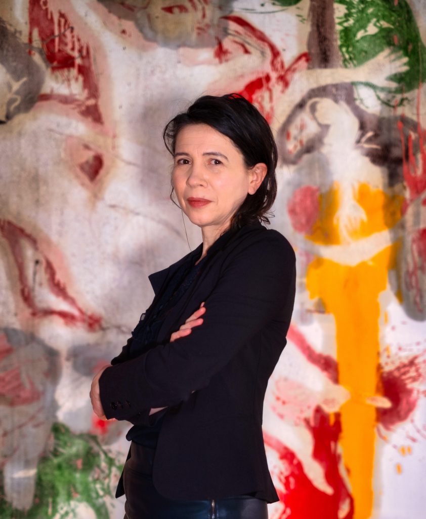 three-quarter length photo portrait of Martina, arms crossed, side view, head turned towards camera, in front of a large semi-abstract vigorously painted image which fills the backgound
