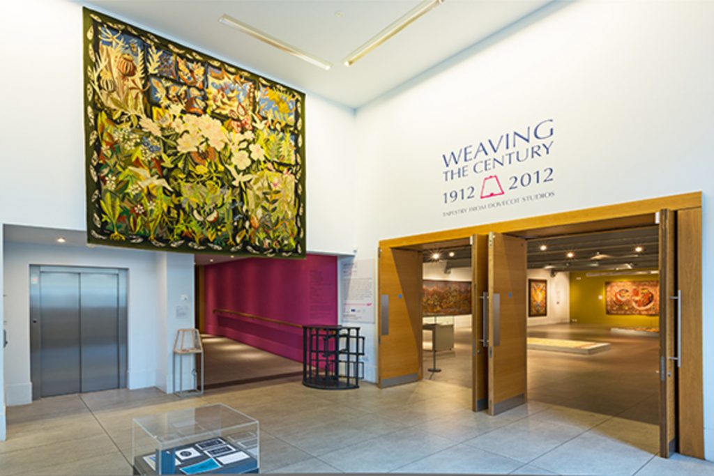 view of the exhibition entrance, a tapestry handing high above a corridor and lift entrance to left, open doorways into the show on the right