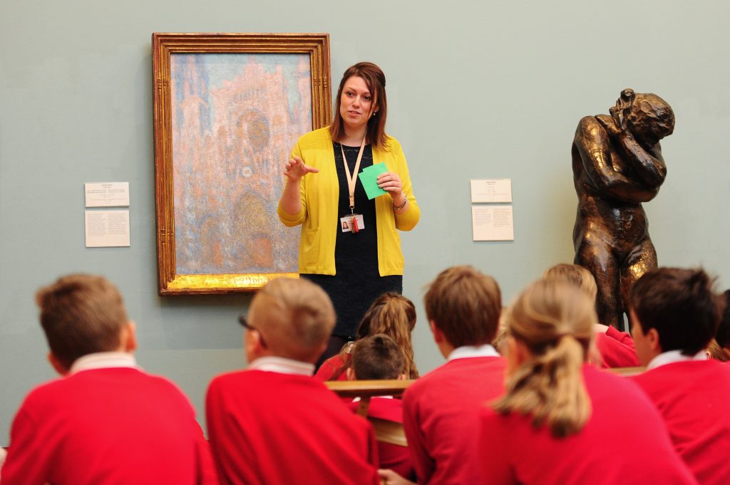 gallery interior - schoolchildren in red tops seen from behind, listening to Steph talk in front of a picture, a sculpture to the right