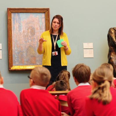 gallery interior - schoolchildren in red tops seen from behind, listening to Steph talk in front of a picture, a sculpture to the right