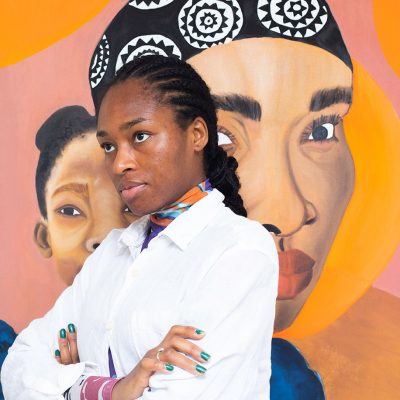 portrait photo of Tobi, looking left out of shot, arms crossed in white shirt, a large scale painting of two faces with abstract pink and orange backgrounds partly obscured behind