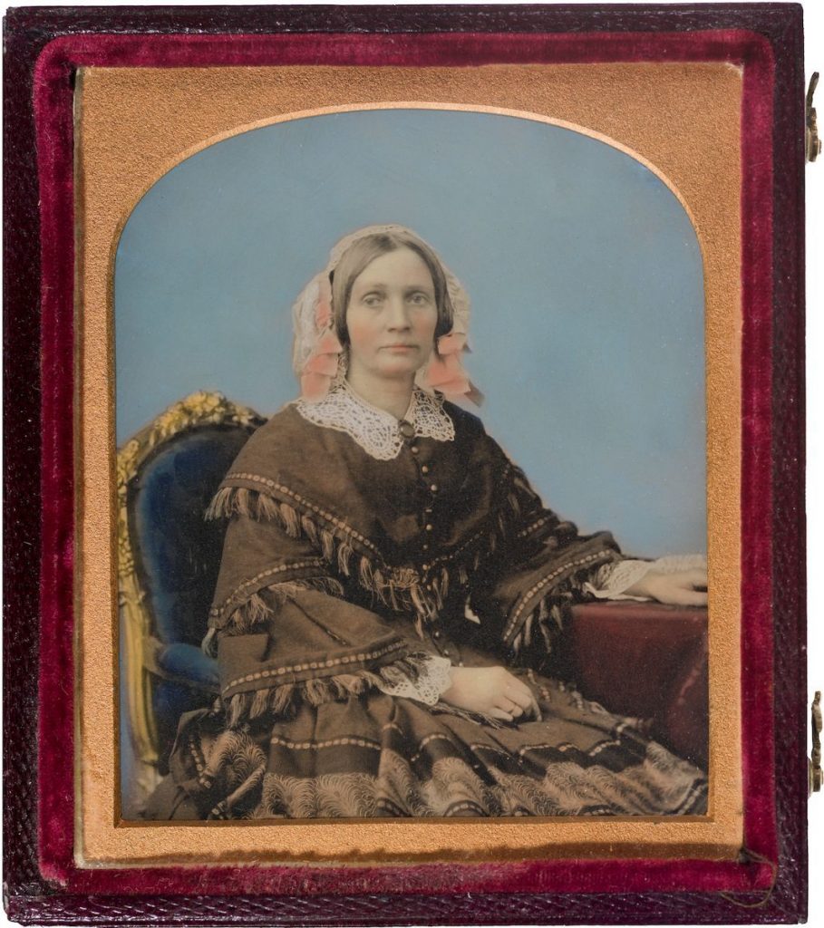 early photographic portrait of a lady looking impassive, seated in fancy victorian costume, features and headdress tinted pink