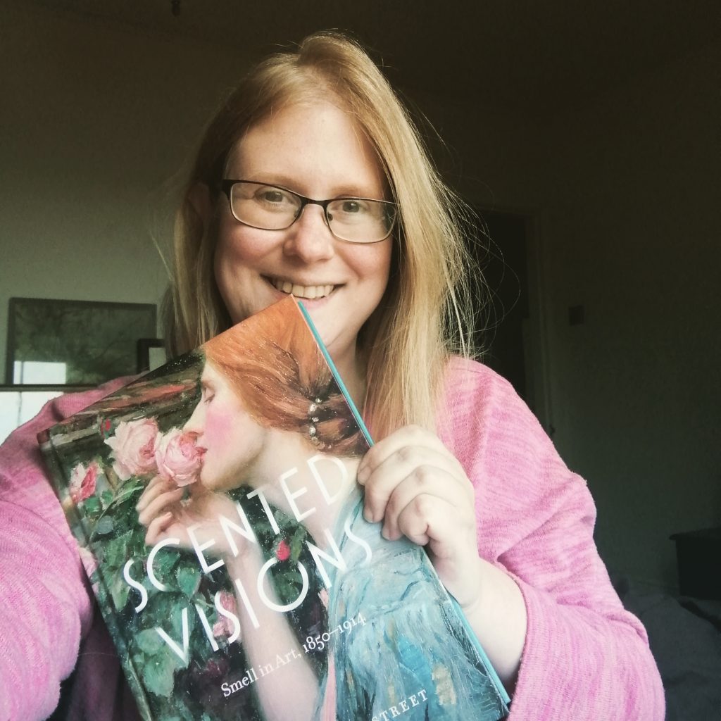 portrait photo of Christina, smiling broadly holding up copy of her book Scented Visions, featuring a detail of a pre-raphaelite style painting