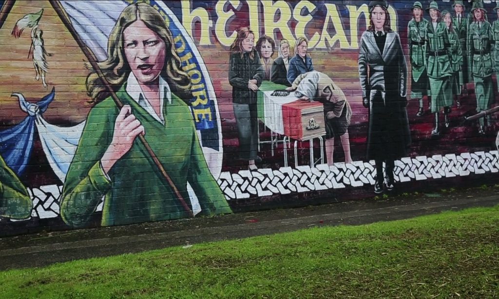 photo of a painted wall mural, a female figure in green to the left of the iamge, a group with a coffin draped in a flag and Celtic lettering to right