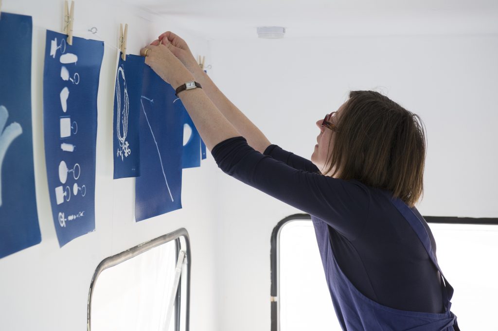 action shot of Jane pegging up a blue unframed artwork alongside other deep blue images on line against a white wall