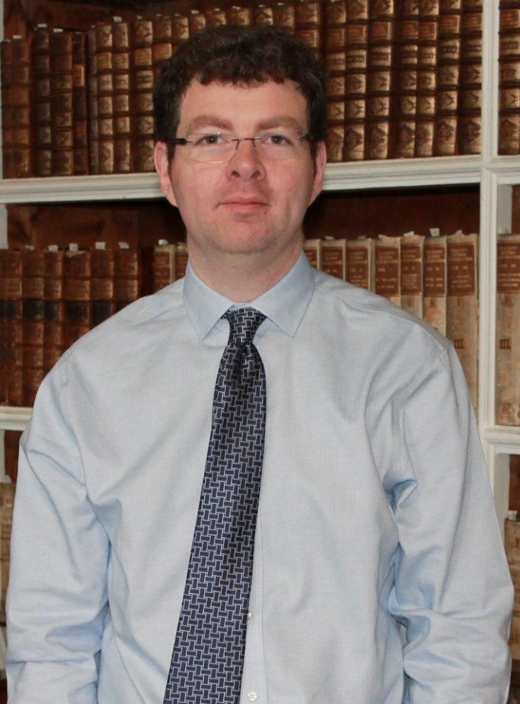 photo portrait of Robert standing in front of shelves packed with historical bound volumes, half length