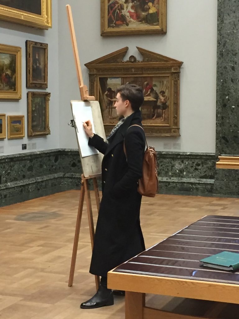photo of Emily standing at an easel in a gallery setting, working on a drawing, PreRaphaelite paintings on walls behind