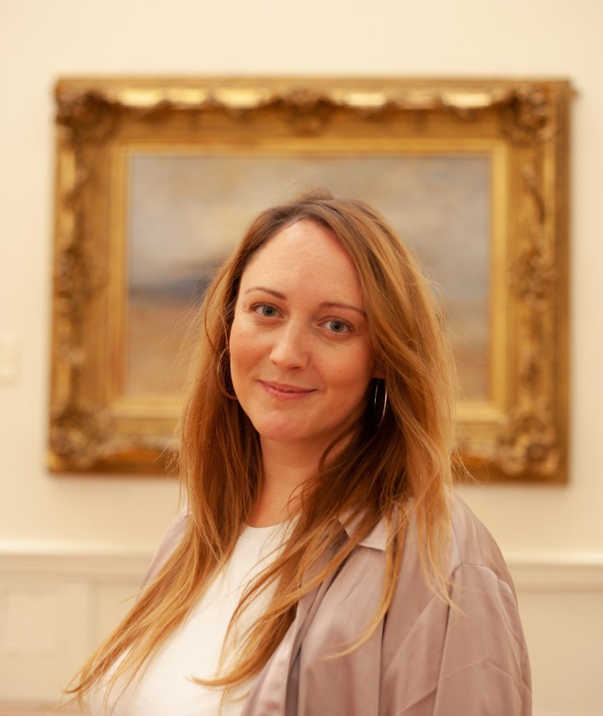 portrait photo of Gina smiling, head and shoulders, in gallery setting, her head framed by a Turner painting on the wall behind