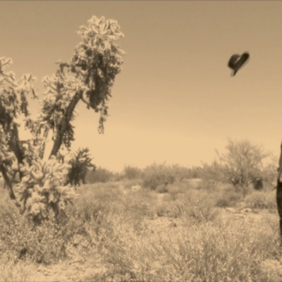 still from video; sepia image of a man looking up at a hat, mid air between himself and a tree, in a barren Arizona desert setting