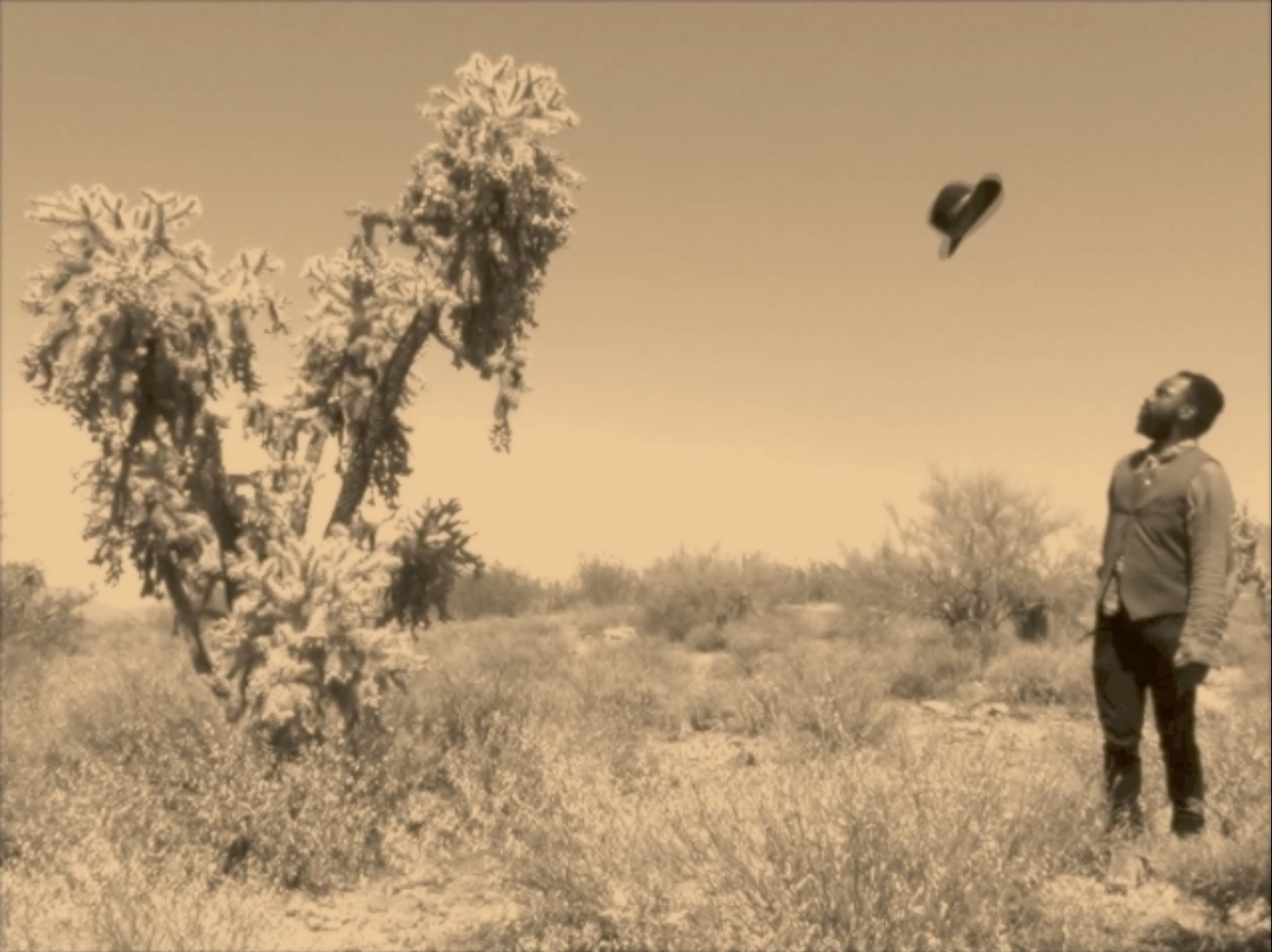 still from video; sepia image of a man looking up at a hat, mid air between himself and a tree, in a barren Arizona desert setting