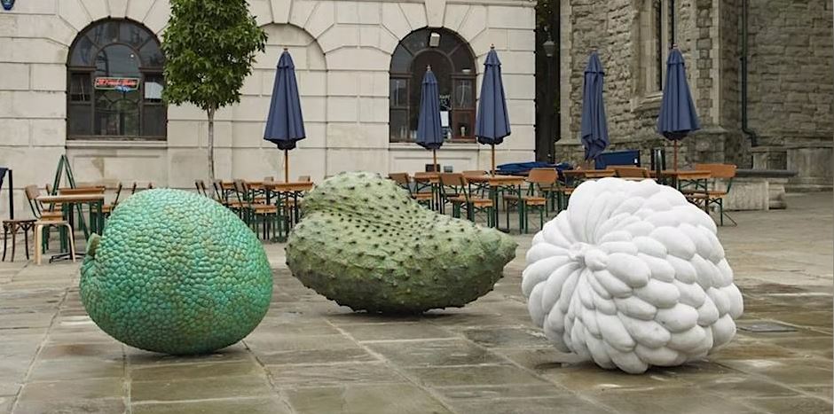 photographic view of a paved square, cafe tables behind, in foreground three sculptural artworks by Veronica Ryan, two green, one white, taking the form of gigantic fruit