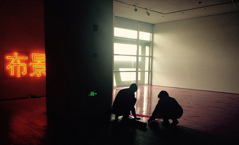a darkened gallery interior, bare red floor, a window behind admitting some daylight, in the foreground two figures in silhouette, crouching; to the left the darkened space is illuminated by a large neon of two Chinese or Japanese characters