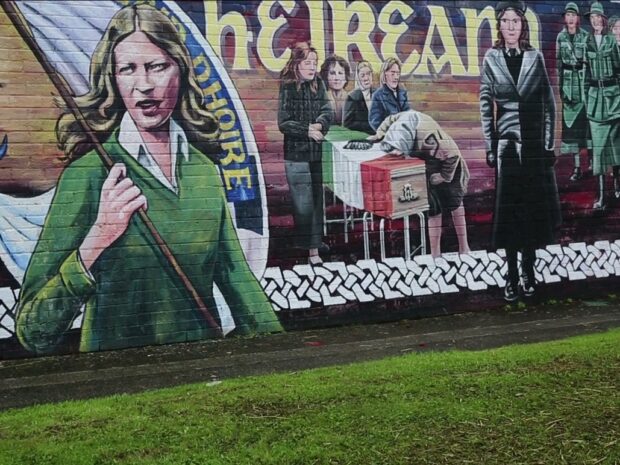 an artists mural - grass before, the mural extending across our view including figures in green military costume, a coffin draped with the Irish flag with mourning figures, and a half-length female figure with a large banner - also lettering in Gaelic