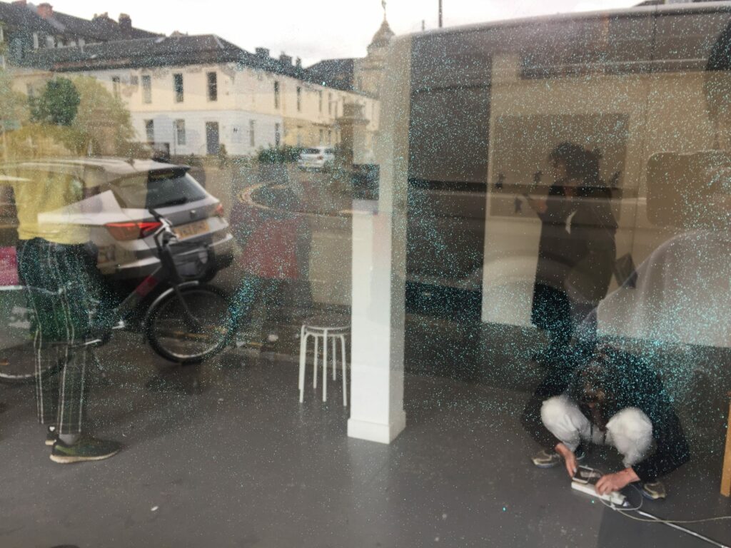 a view through glass into a gallery interior, with four or more figures indistinctly seen crouching or standing - their figures combine with the reflected view of a street with cars, bikes and buildings