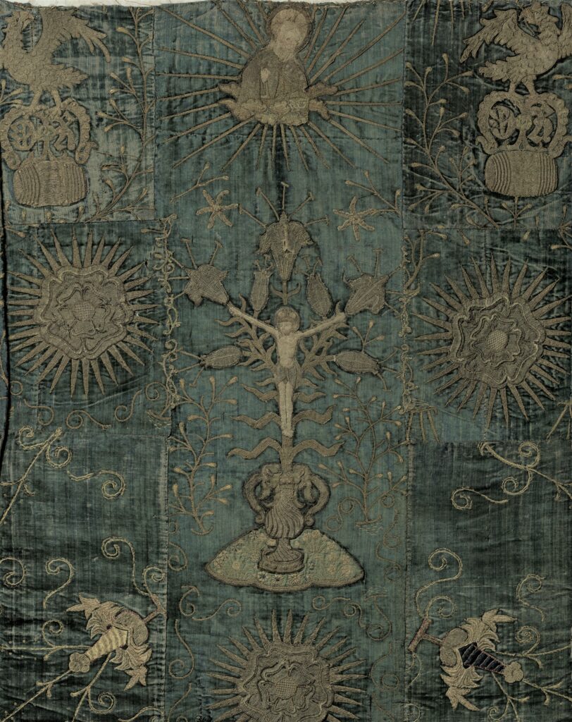 close up of a tapestry in deep greens and browns, featuring Christian religious imagery including flowers and a crucified Christ