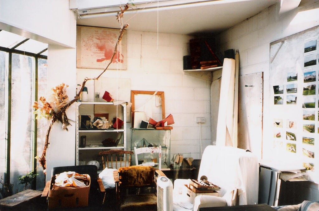 an interior, an artist's studio, bare white painted walls, a window to the left, with a branch suspended before it, various furniture crowded together on the floor, a wall of photographic images in colour of landscape scenes pinned up to the right - a large rolled canvas to the rear, leaning against a shelf