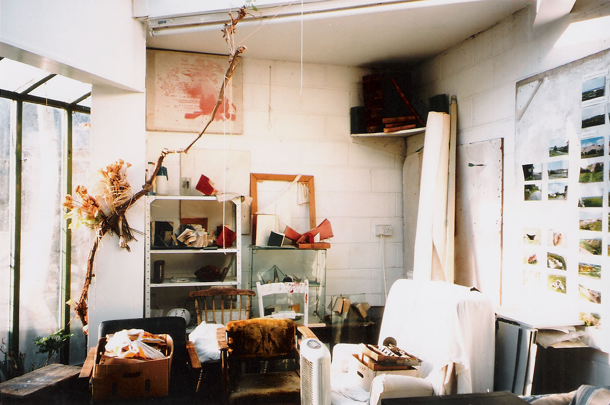 an interior, an artist's studio, bare white painted walls, a window to the left, with a branch suspended before it, various furniture crowded together on the floor, a wall of photographic images in colour of landscape scenes pinned up to the right - a large rolled canvas to the rear, leaning against a shelf