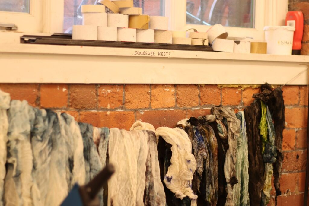 an assortment of cloths stained with different coloured inks hang on a line below an interior window