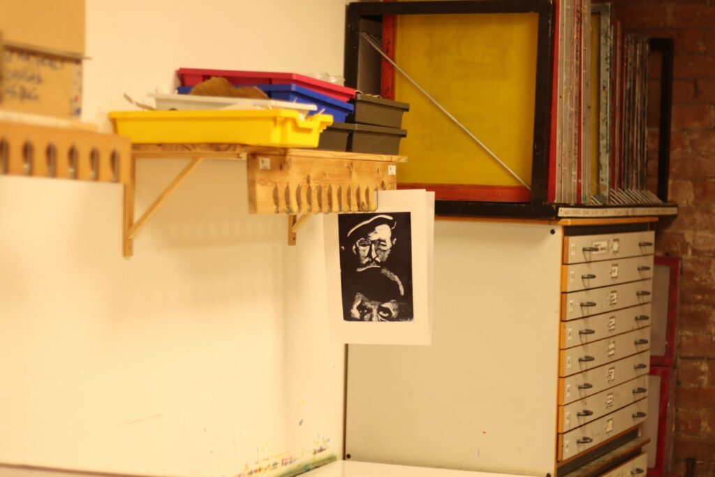 the inside of a print studio with drawers and screens with mesh. A black and white print depicting two faces hangs in the centre.