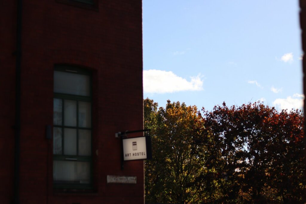 the corner of a red brick building in shadow with a sign reading 'art hostel'. to the left are the upper branches and leaves of a tree