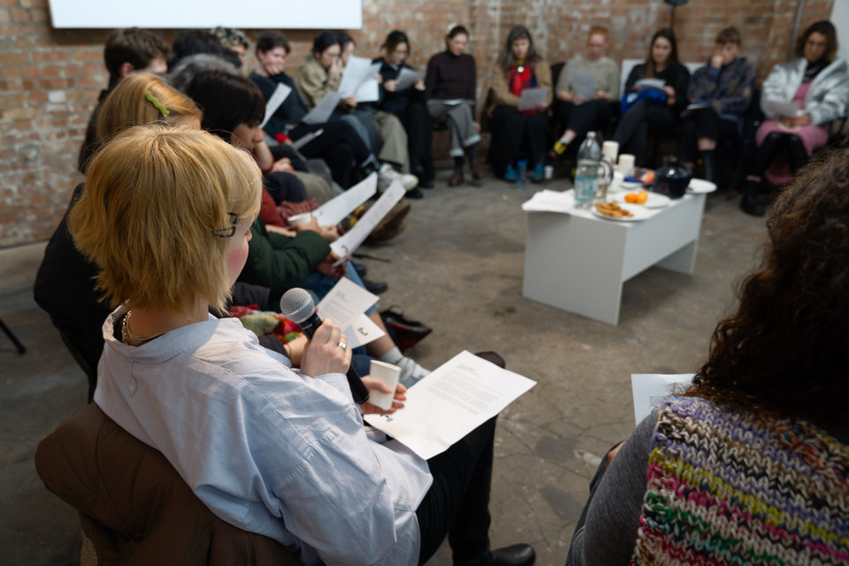 A woman with a microphone speaks to a group of people in a circle during an indoor workshop.