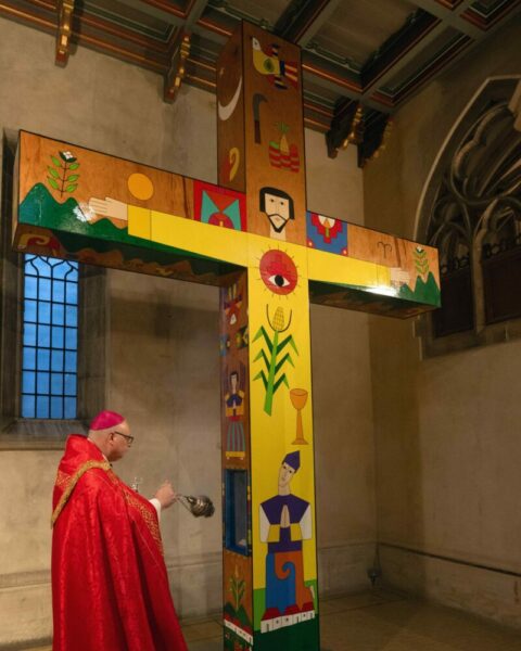 A priest wearing red robes blesses a large, brightly painted crucifix