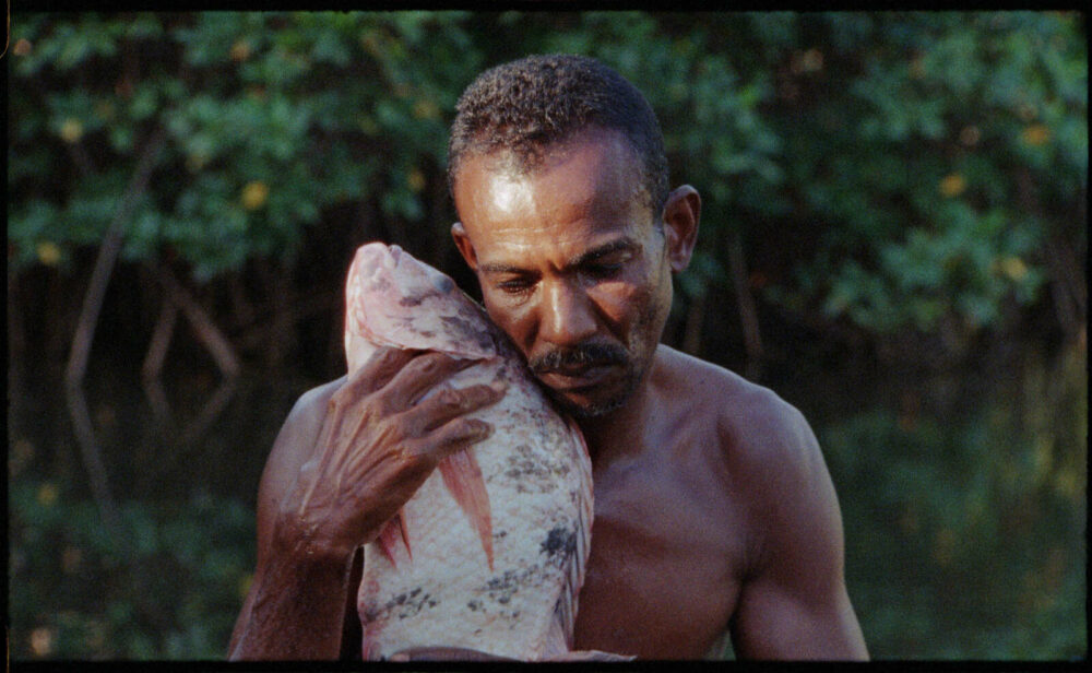 Close up of a shirtless man cradling a large fish to his face. In the background are trees and bushes.
