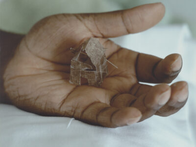 A close up image of an open hand holding a very small, fragile sculpture of a house. The house is made from a thin, brittle fibre and is held together with two pins.