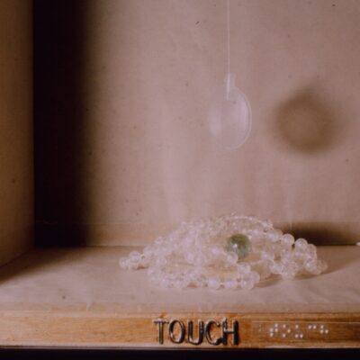 A wooden box containing a string of glass beads, a magnifying glass and a marble. The lower edge of the box has the word "Touch" in capital letter and in braille.